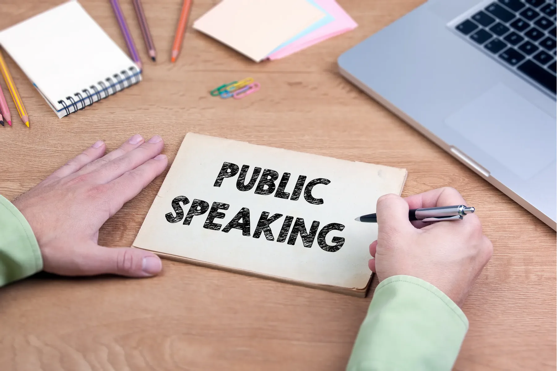 A person at a desk writing the words public speaking on a card, with notebooks, pencils and a laptop nearby.