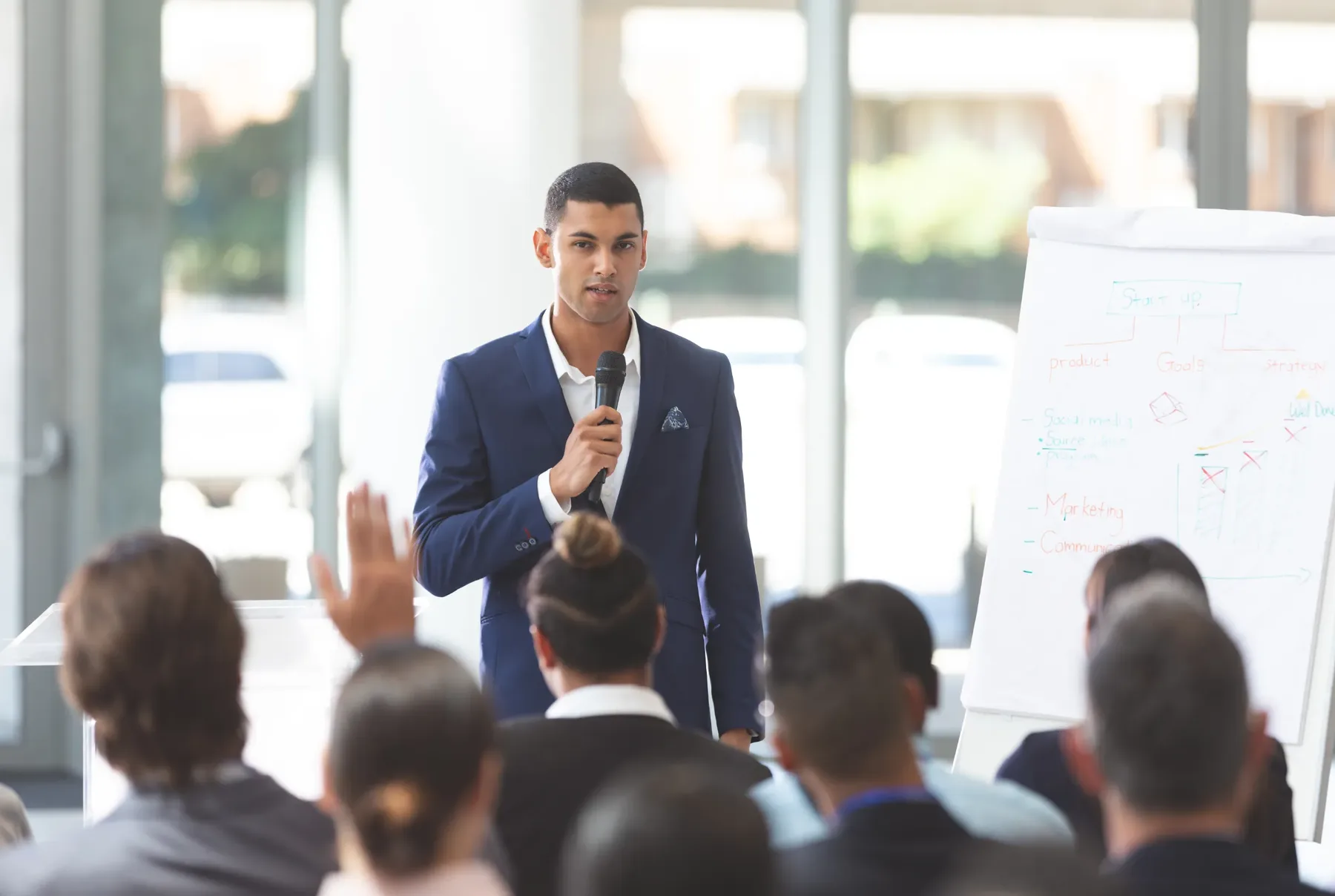A man in a navy suit speaking into a microphone in front of a seated audience, with a flip chart beside him.