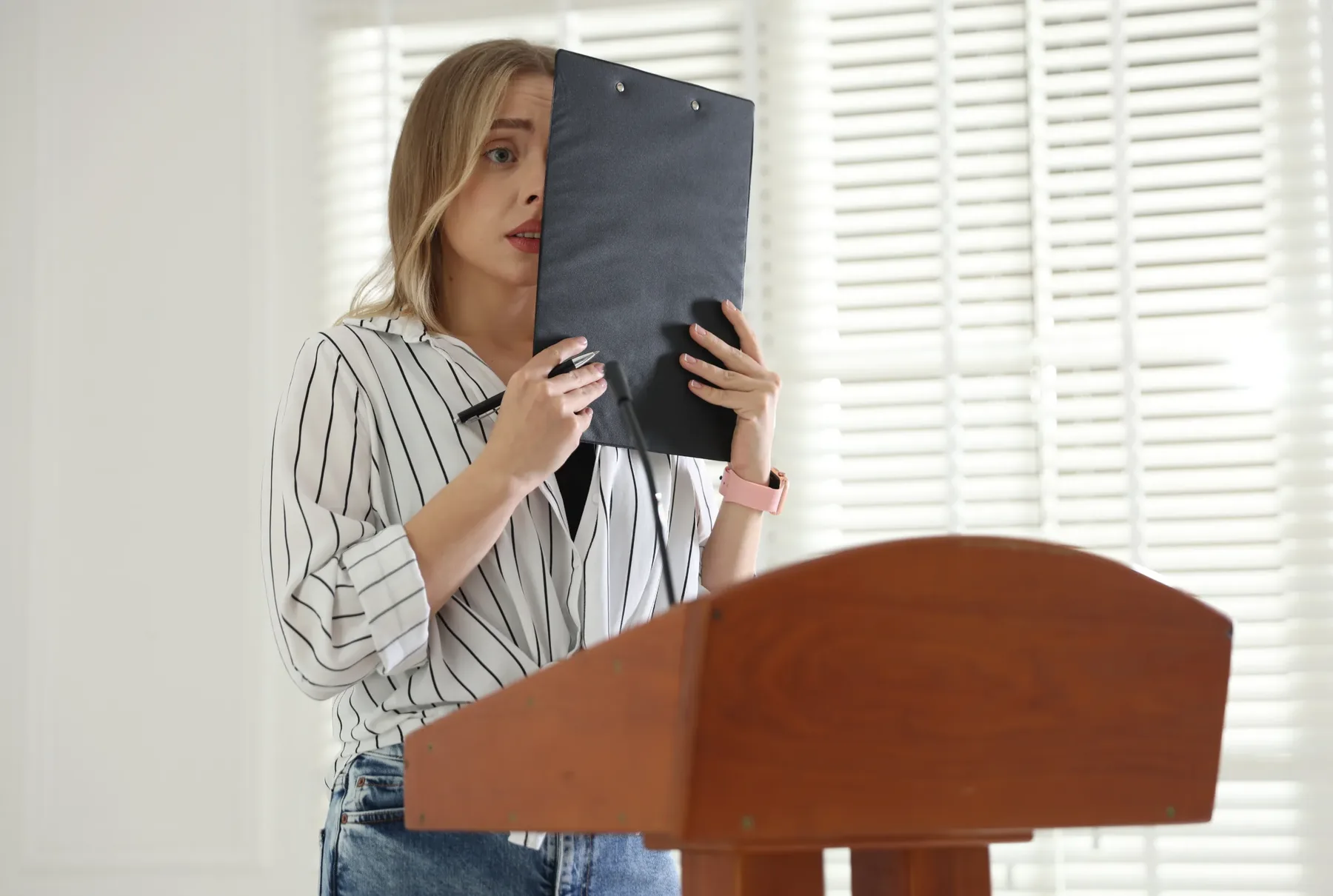 A woman at a lectern holding a clipboard up in front of her face, appearing apprehensive before speaking.