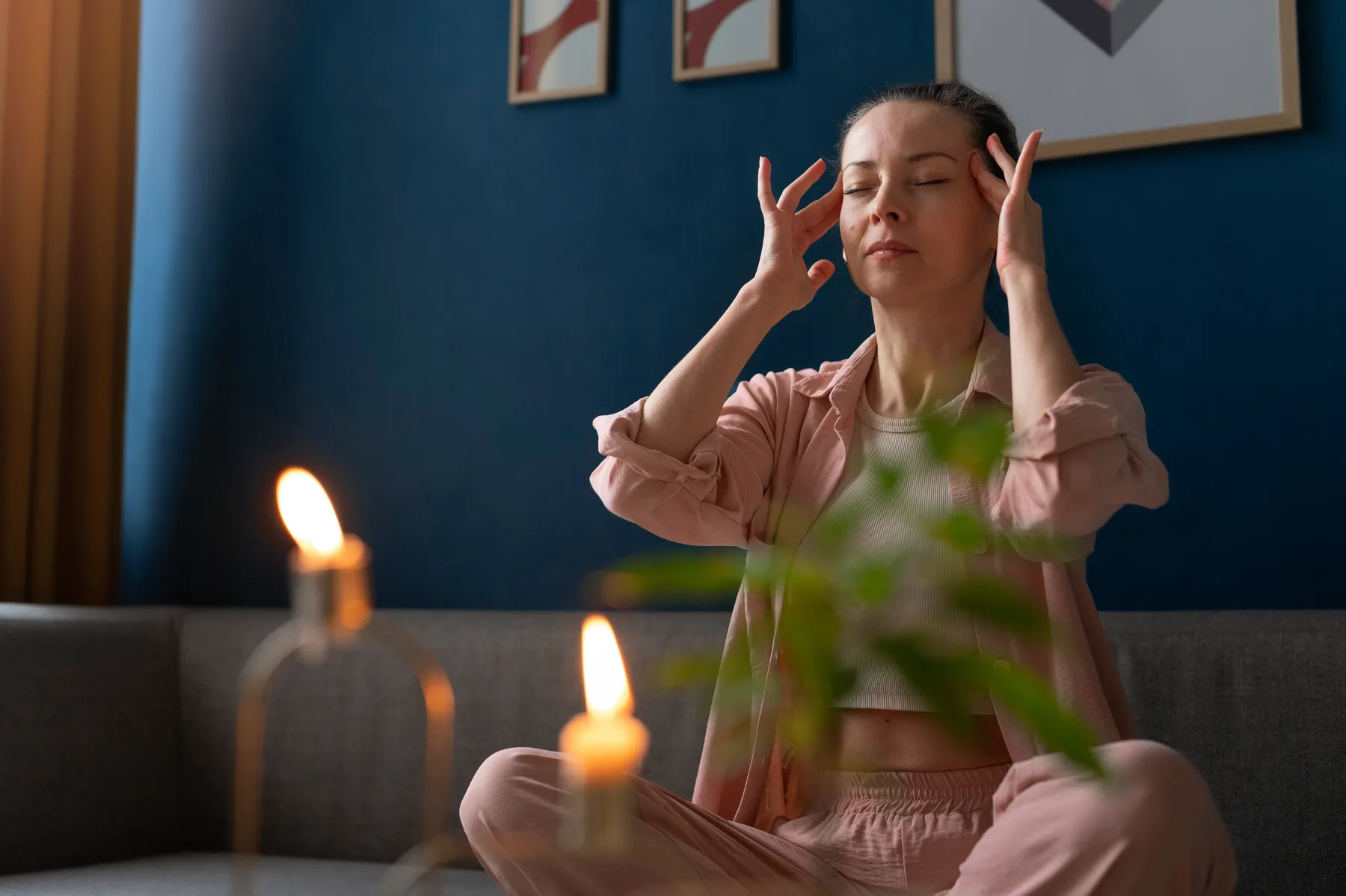 A person sitting cross-legged with eyes closed and fingertips at their temples, in a quiet room lit by candles.