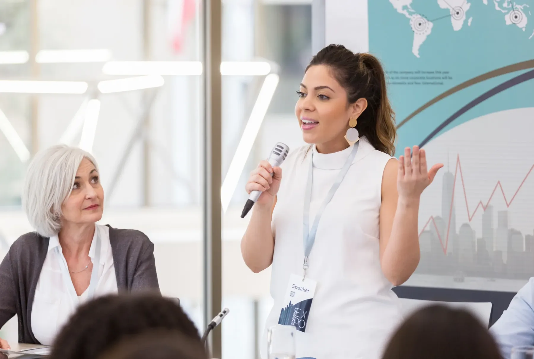 A professional speaking into a microphone at a conference table while a colleague listens attentively.